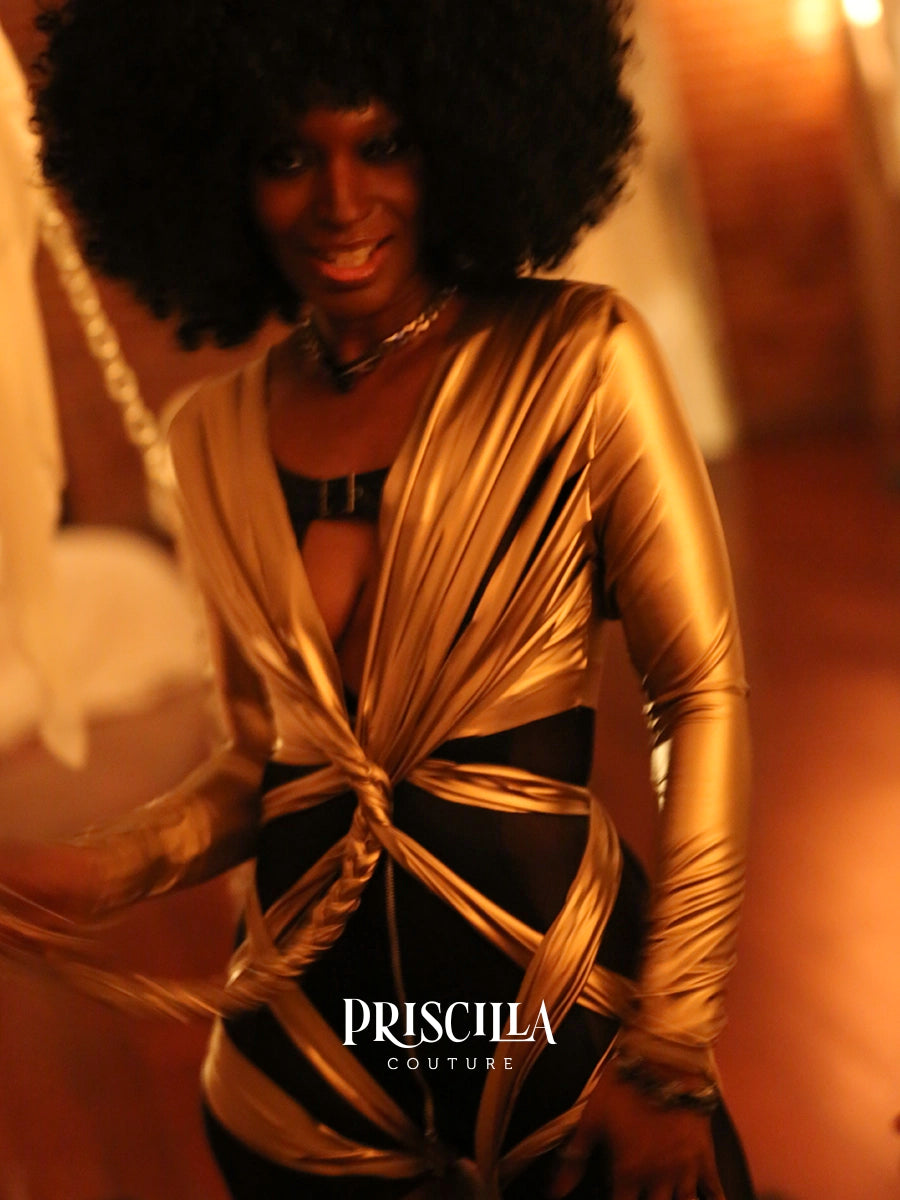 A woman wearing a metallic gold wrap-style lingerie with black underlayer, photographed in warm orange-toned lighting. The brand name PRISCILLA COUTURE is visible at the bottom.