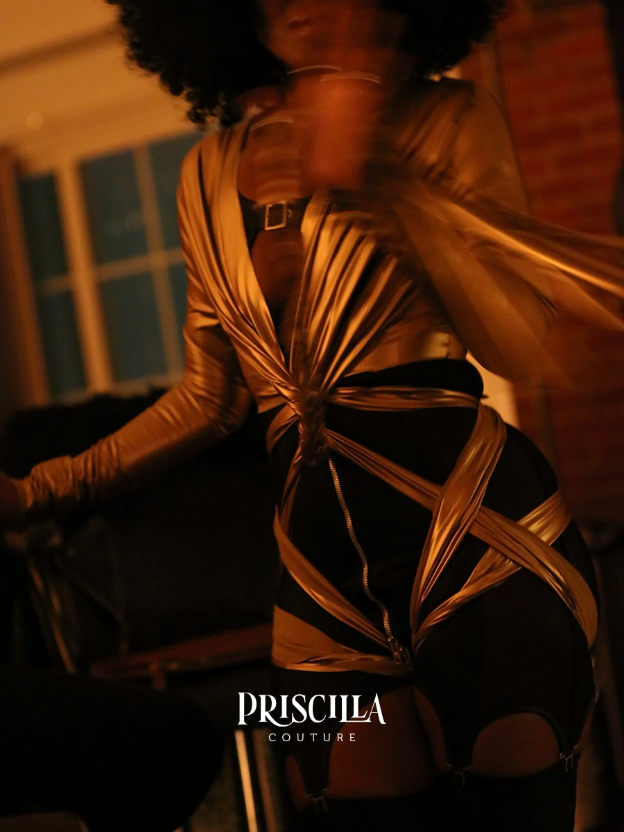 A woman wearing a metallic gold draped lingerie with black strappy detailing over black bottoms, photographed in warm dim lighting with a blurred indoor background. The brand name PRISCILLA COUTURE is visible at the bottom.
