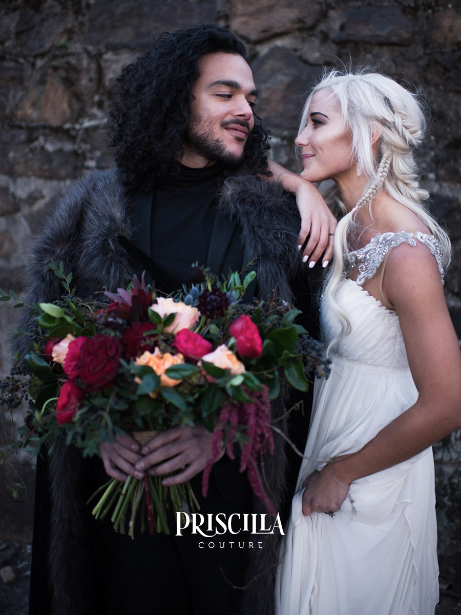 A couple in wedding attire standing together against a rustic stone wall. The man wears a black outfit with a dark fur-look coat and holds a large bouquet of red, pink, and peach flowers. Woman is dressed in a Priscilla Couture sleeveless white wedding dress with silver shoulder strap embellishment.