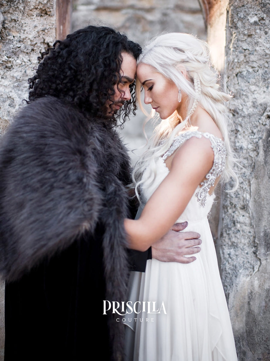A bride in a white lace and silk wedding gown embraces a groom wearing a black coat with a dark gray fur collar, posed against a rustic stone wall. The brand name 'PRISCILLA COUTURE' is watermarked at the bottom.