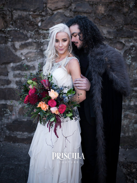A bride in a white silk chiffon wedding dress holding a red and orange floral bouquet stands with a groom in a dark fur-trimmed cloak against a rustic stone wall. The brand name 'PRISCILLA COUTURE' is watermarked at the bottom.