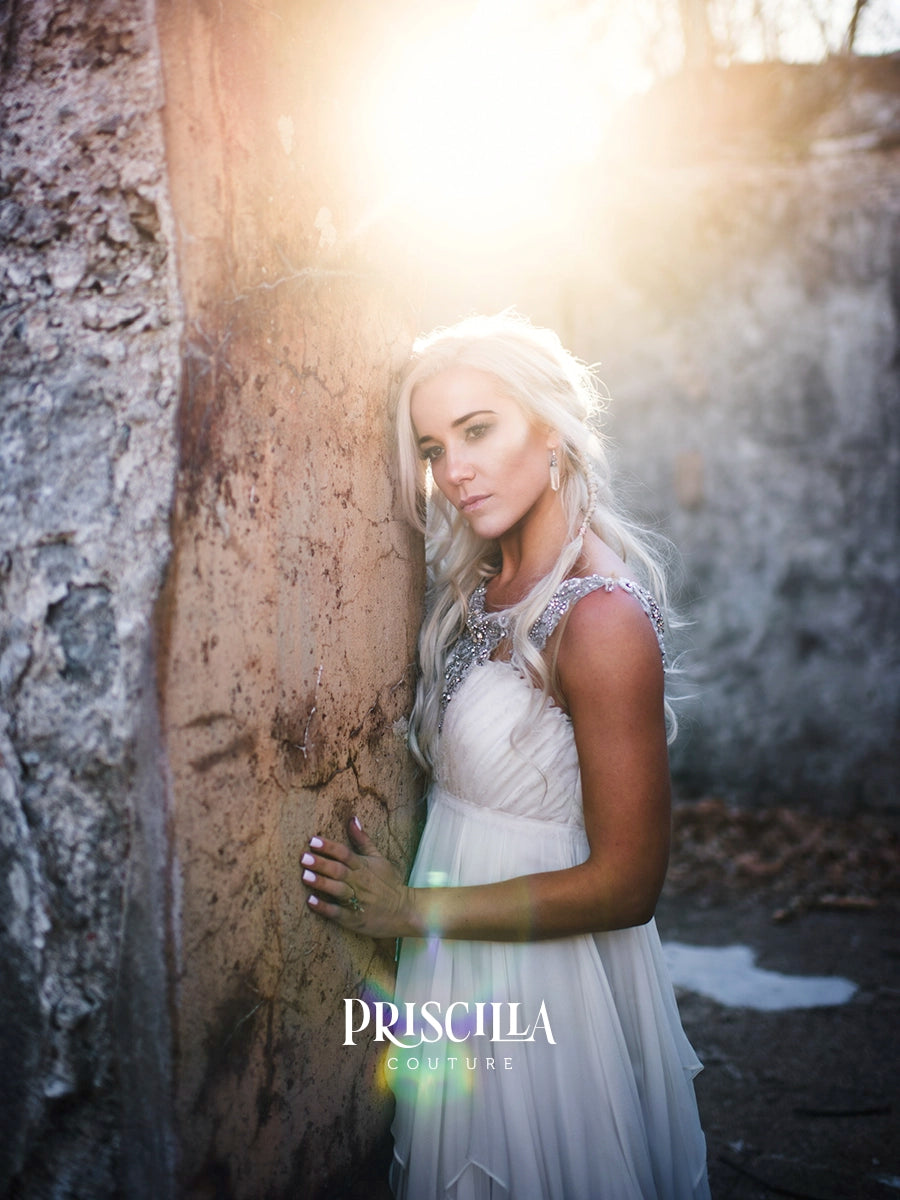 A woman with long platinum blonde hair stands outdoors against a textured stone wall, wearing a white sleeveless dress with embellished shoulders. Strong backlighting creates a bright glow, and the brand name 'PRISCILLA COUTURE' appears at the bottom.