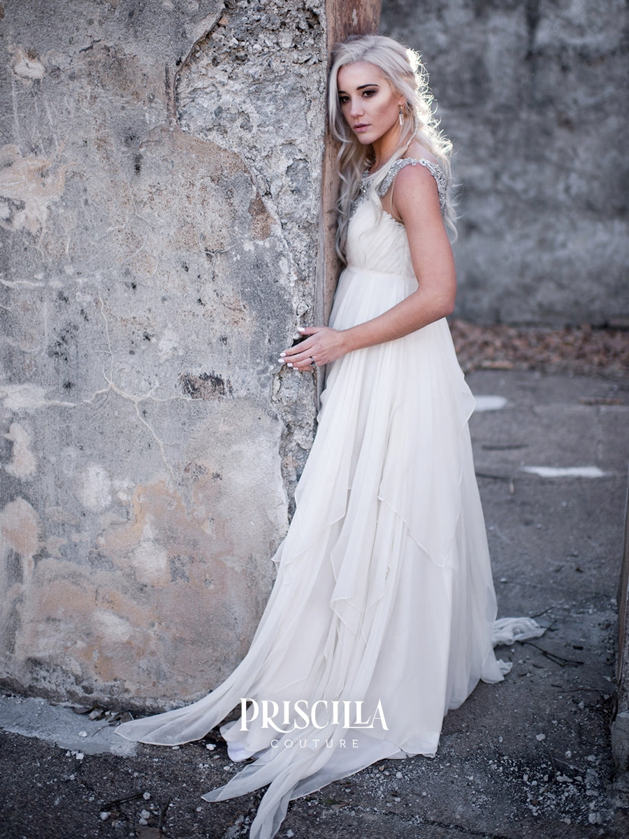 A woman wearing a flowing white silk chiffon wedding dress with sheer cap sleeves and a long layered skirt, leaning against a weathered stone wall outdoors. The brand name PRISCILLA COUTURE is printed at the bottom.