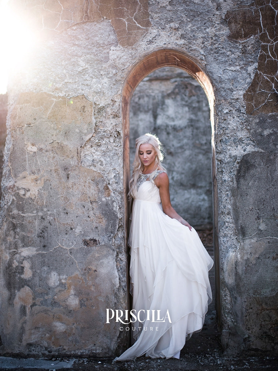 A woman in a Priscilla Couture white silk chiffon wedding gown stands in a rustic stone archway, with bright sunlight streaming from the upper left. The dress is a sleeveless, tiered A-line gown with a sheer lace bodice and a full layered skirt.