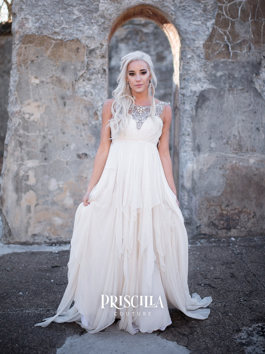 A woman wearing a flowing ivory Priscilla Couture silk wedding gown stands in front of a rustic stone archway. The dress has a sheer embellished neckline and a full layered skirt with a train.