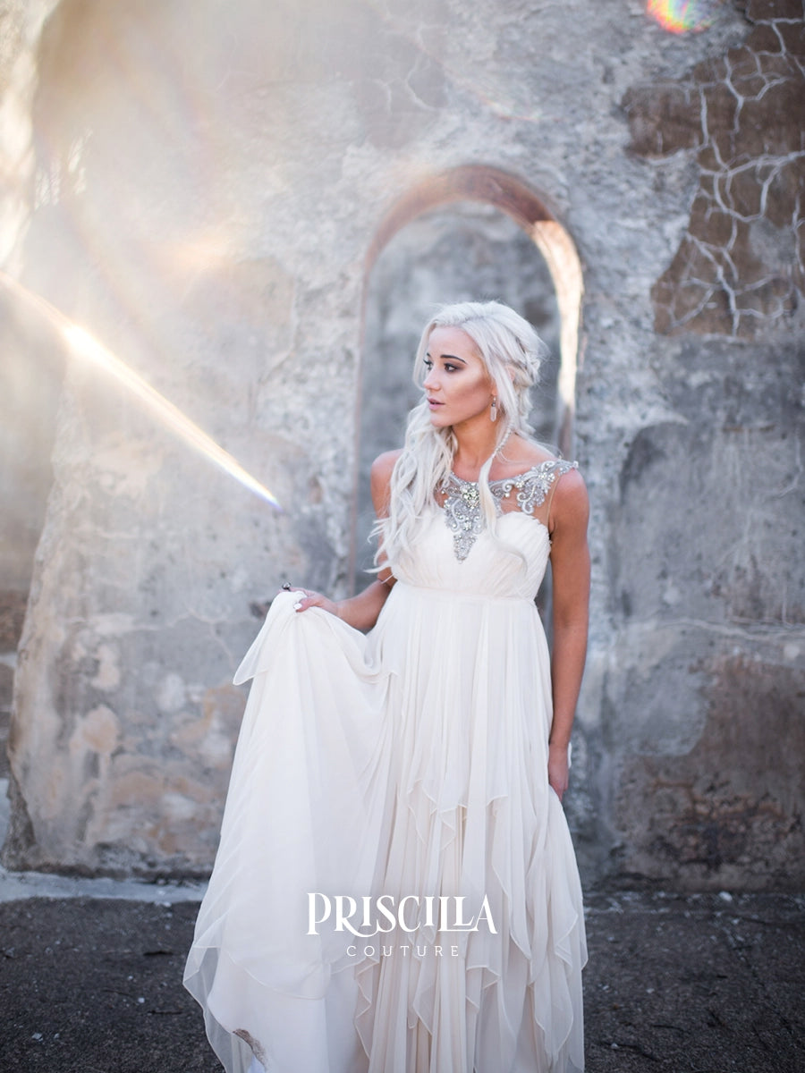 A woman wearing a white Priscilla Couture wedding gown with silver embellished straps and a flowing tiered skirt, standing in front of a rustic stone wall with dramatic sunlight and lens flare.