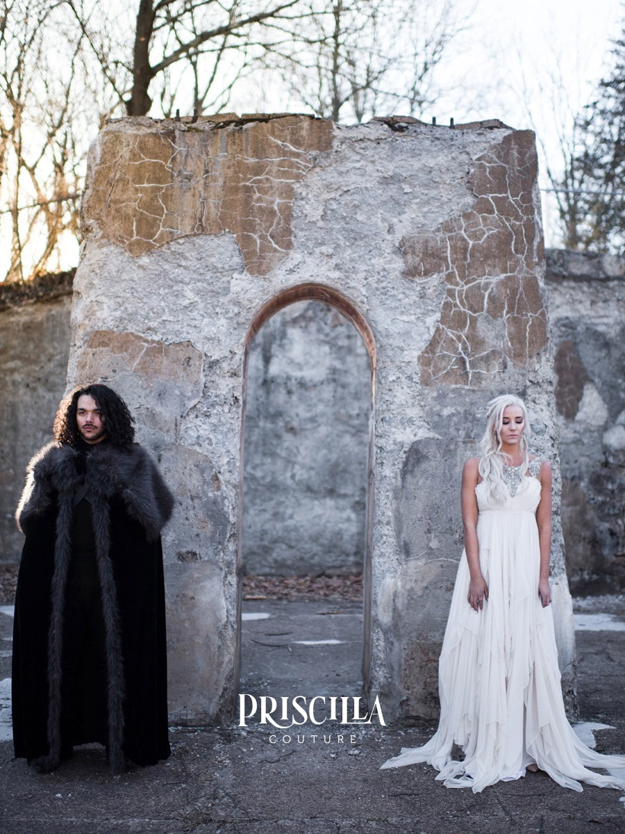 A man in a long black fur-trimmed cloak stands on the left, and a woman in a flowing white silk gown stands on the right, posed in front of a weathered stone ruin with bare trees in the background. The brand name 'PRISCILLA COUTURE' is printed at the bottom center.