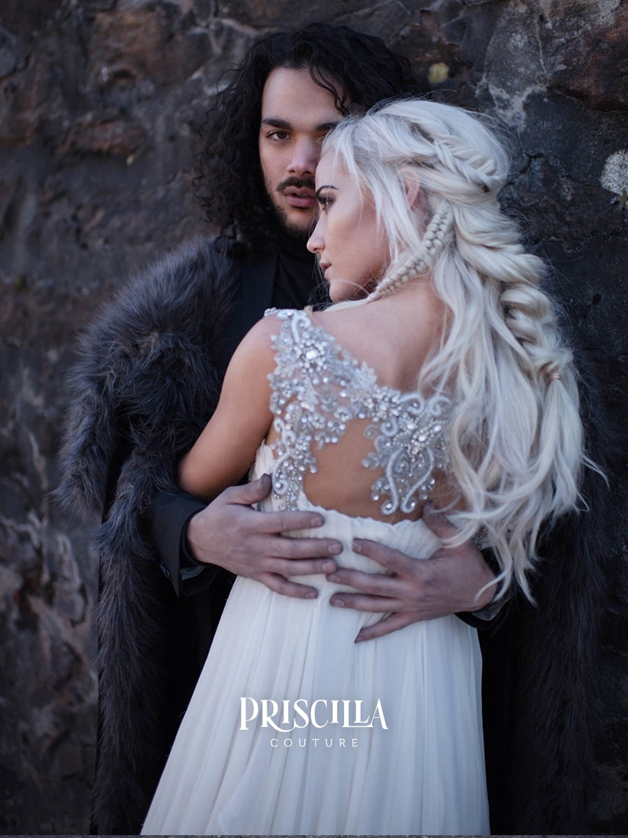 A woman in a white bridal gown with an ornate beaded back and long platinum-blonde hair is embraced by a man in a dark fur-lined cloak against a rocky background. The brand name PRISCILLA COUTURE is printed at the bottom.