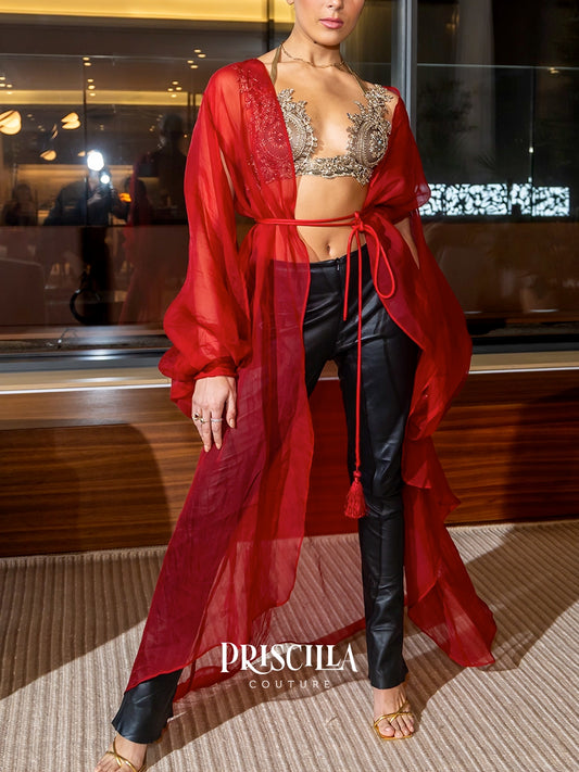 A model wearing a vibrant red sheer long-sleeve drape robe over a silver embellished bralette and black faux-leather pants, standing in an indoor setting with a 'PRISCILLA COUTURE' watermark at the bottom.