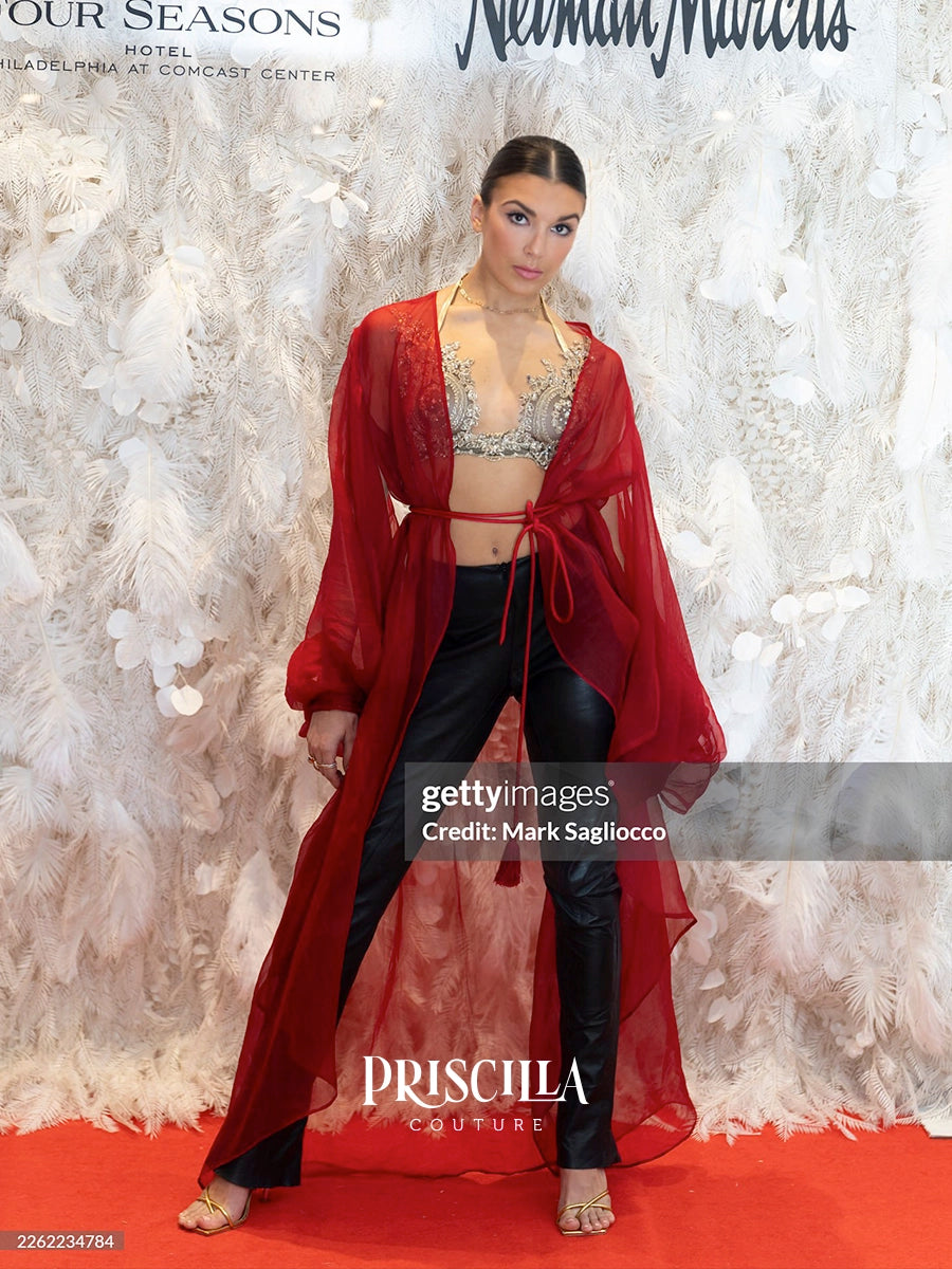 Priscilla Couture model posing on a red carpet in front of a white feathered backdrop, wearing a sheer red open-front robe over a silver embellished crop top and black pants.