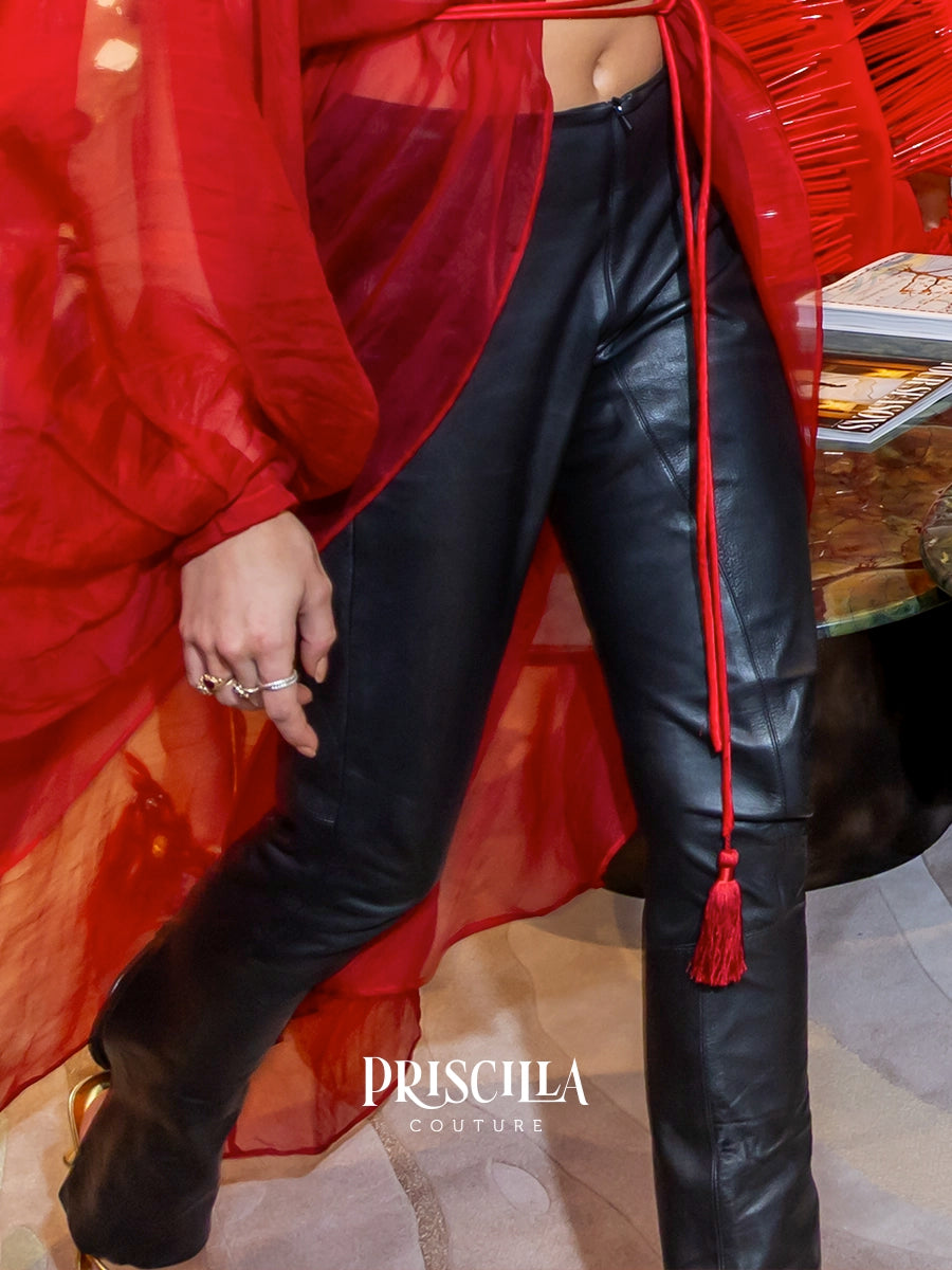 A person wearing black leather pants and a sheer red wrap top, with a red tassel drawstring hanging down. The brand name 'PRISCILLA COUTURE' is watermarked at the bottom.
