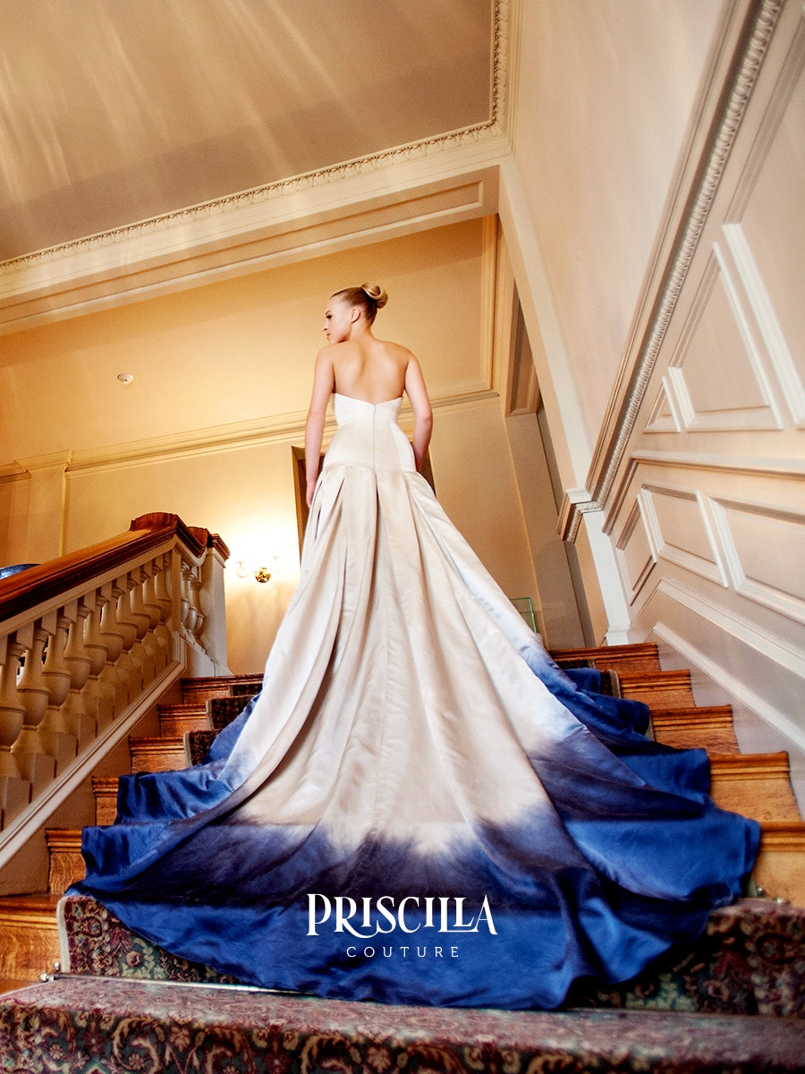A woman wearing a Priscilla Couture silk duchesse A-line strapless colored bridal gown stands on an indoor staircase, facing away from the camera. The dress has a cream upper section that fades into a deep blue ombré at the bottom.