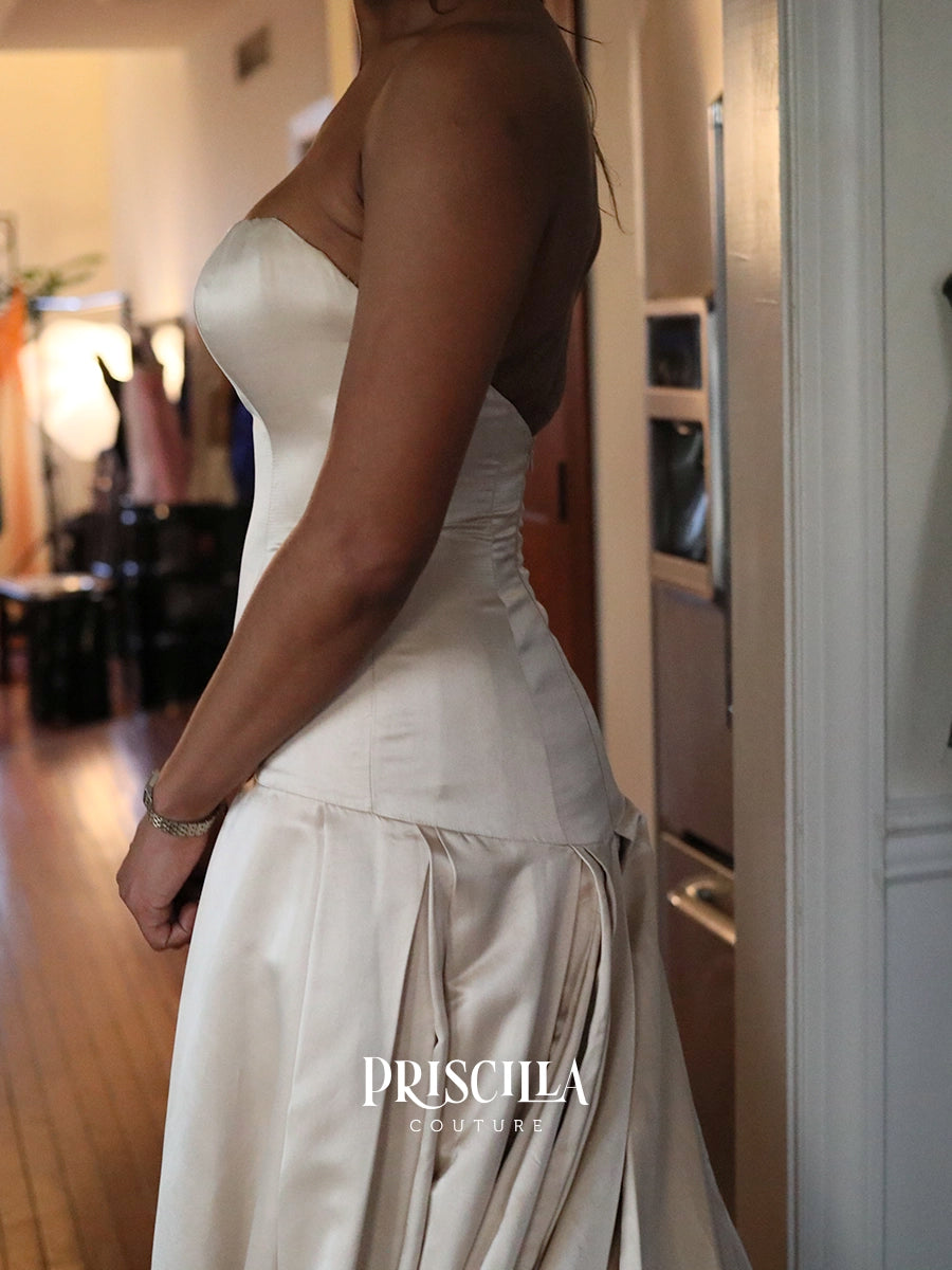A woman wearing a strapless ivory silk duchesse wedding gown with a structured corset-style bodice and draped skirt, photographed indoors in a home setting. The brand name 'PRISCILLA COUTURE' is watermarked at the bottom.