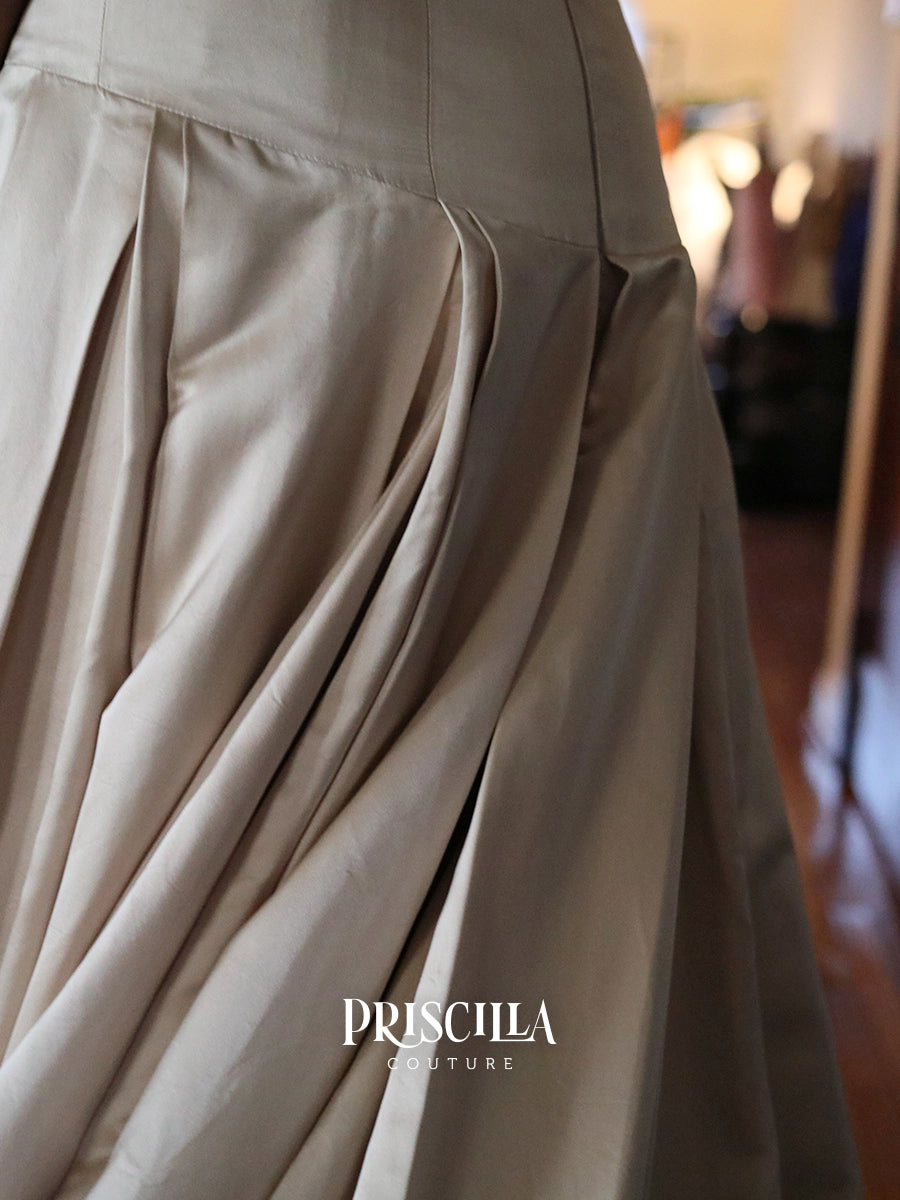 Close-up of a pleated champaign skirt with a satin-like sheen, featuring the Priscilla Couture logo at the bottom.