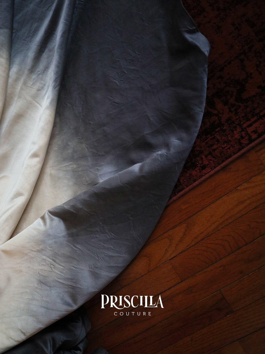 A draped piece of Priscilla Couture wedding dress train with a metal gun grey to off-white ombré finish, shown on a wooden floor next to a dark patterned rug.