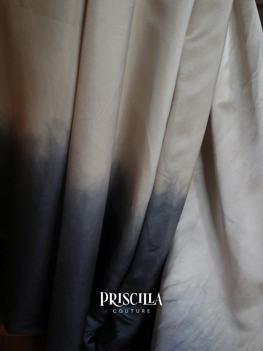 A close-up of wedding dress fabric with a vertical ombré transition from warm beige at the top to dark charcoal gray at the bottom. The brand name 'PRISCILLA COUTURE' is printed in white text near the bottom center.