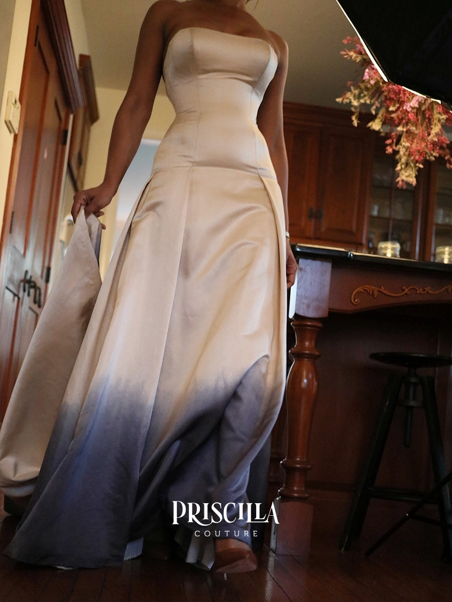 A strapless floor-length silk duchesse gown worn by a model in an indoor room with wood floors and cabinetry. The dress has a fitted bodice and a flowing skirt with a smooth gradient from warm ivory at the top to slate blue-gray at the bottom.