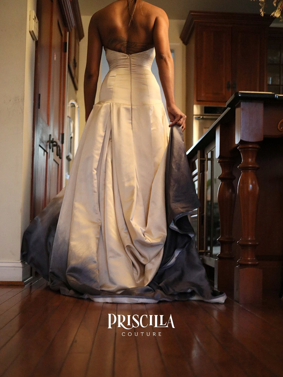 A woman viewed from behind wearing a floor-length bridal gown with a structured ivory bodice and a dark gray satin overskirt, standing on a wooden floor in a home interior. The brand name 'PRISCILLA COUTURE' is printed at the bottom.