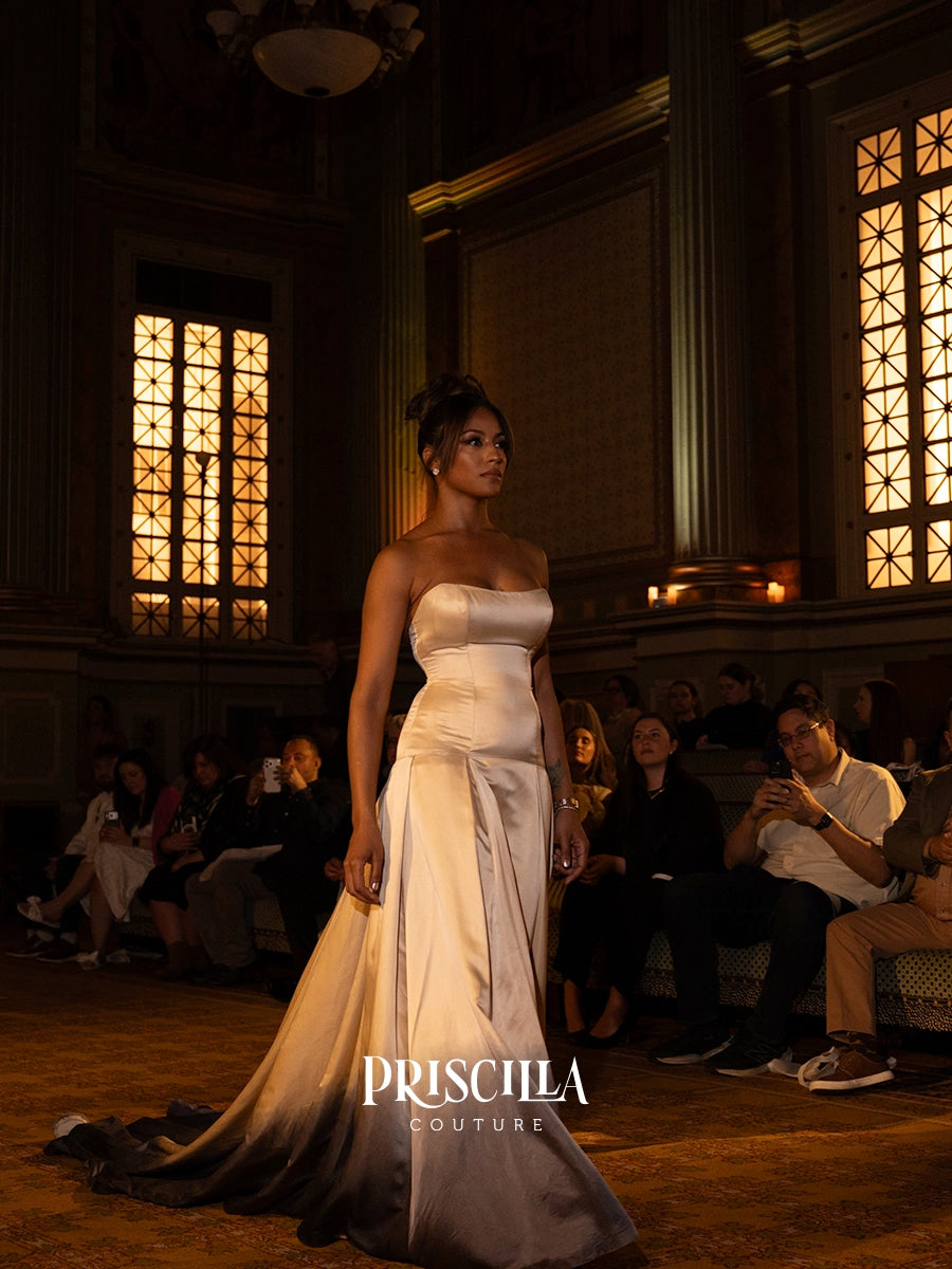A model walks the runway wearing a strapless white wedding gown with a satin bodice and a flowing satin overskirt that fades into a dark train. The event takes place in a grand, dimly lit hall with seated audience members and warm-toned windows in the background.