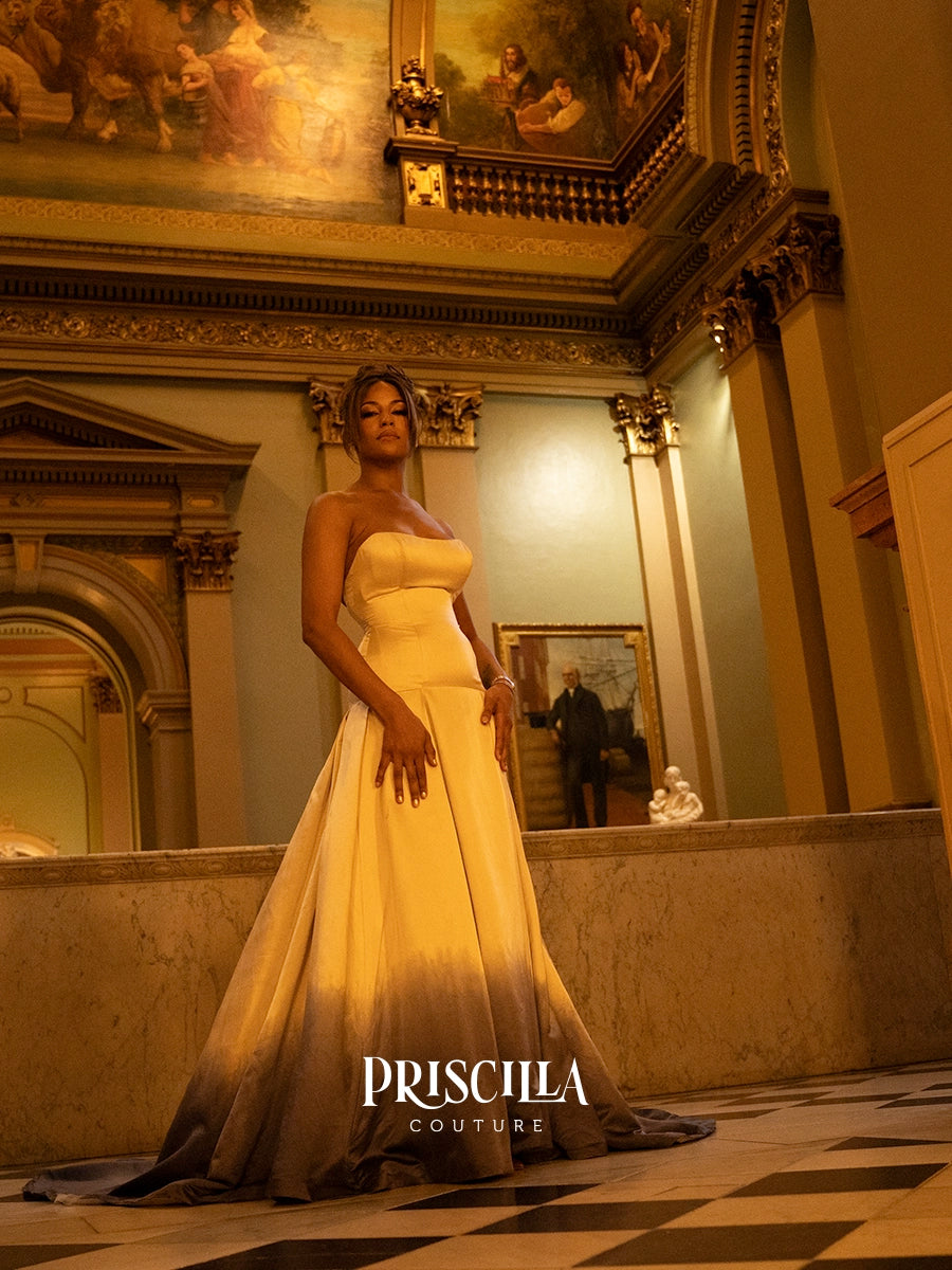 A woman wearing a strapless A-line gown with a fitted bodice and a dramatic gradient hem, standing in an ornate historic interior hall. The gown is cream at the top fading to a dark charcoal/black at the bottom.