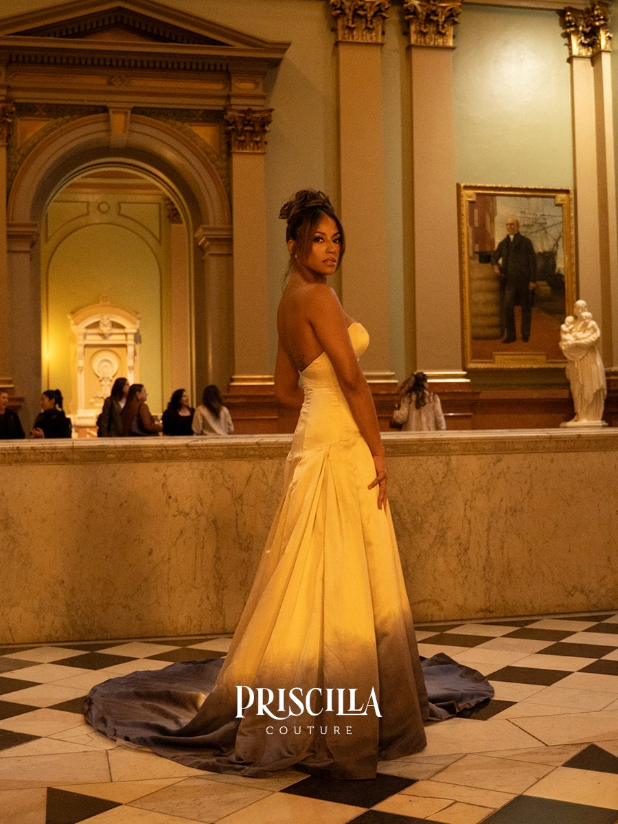 A woman wearing a strapless champagne-colored ball gown stands in a grand indoor hall with classical architecture, a checkered floor, and a visible PRISCILLA COUTURE watermark at the bottom.