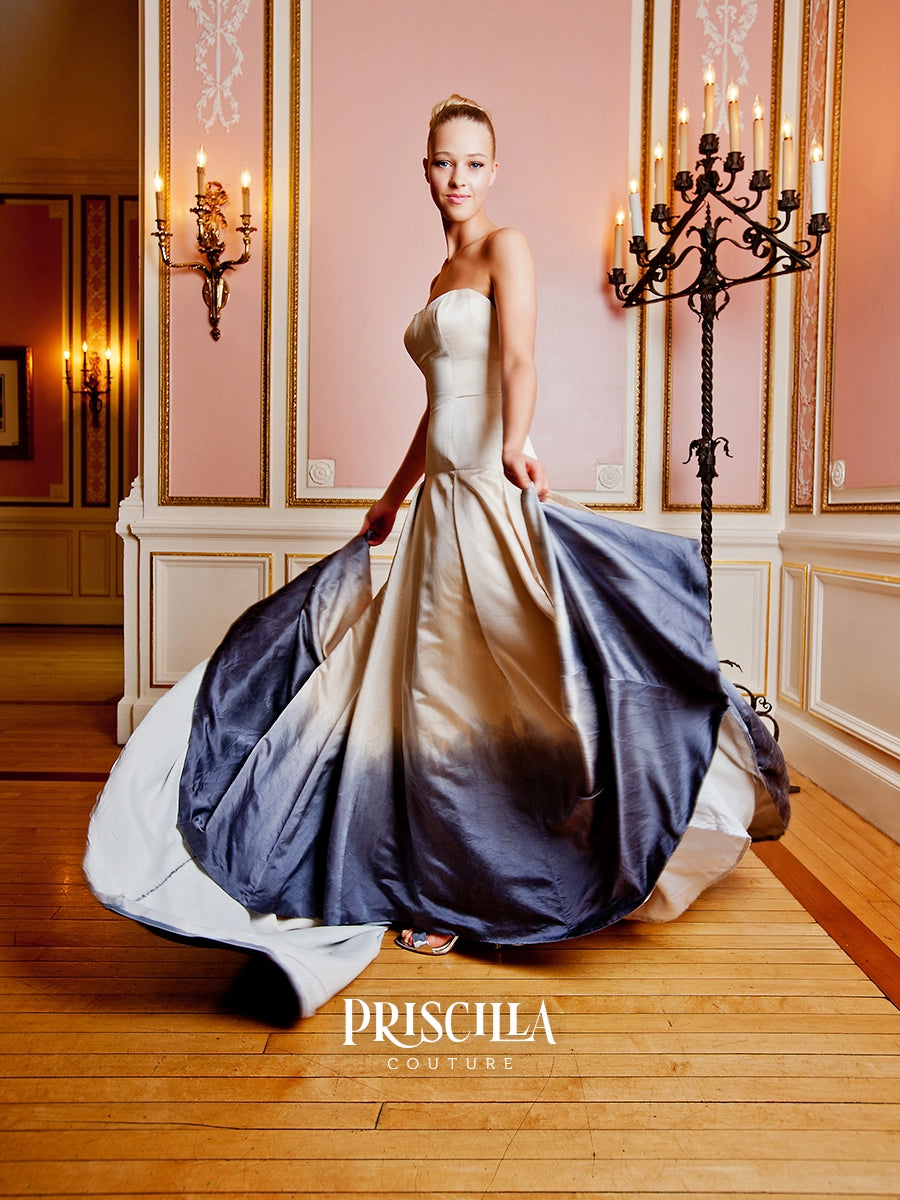 A woman models a strapless silk duchesse A-line wedding gown with a  dramatic navy blue to white ombré satin train, posed in an elegant room with pink walls and candelabras. The brand name 'PRISCILLA COUTURE' is displayed at the bottom.