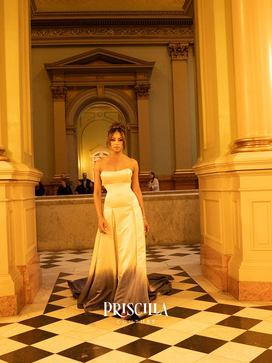 A model stands in a grand, warm-toned hall wearing a strapless white gown with a dramatic gradient hem that fades into black. The background features ornate columns, arches, and a black-and-white checkered floor.