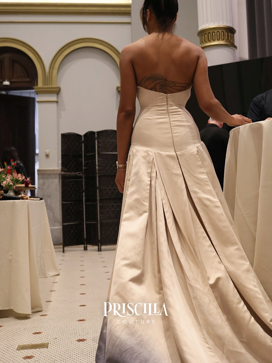 A woman viewed from behind wearing a strapless satin wedding gown with a structured back and a long train in a grand indoor venue. The brand name 'PRISCILLA COUTURE' is watermarked at the bottom.
