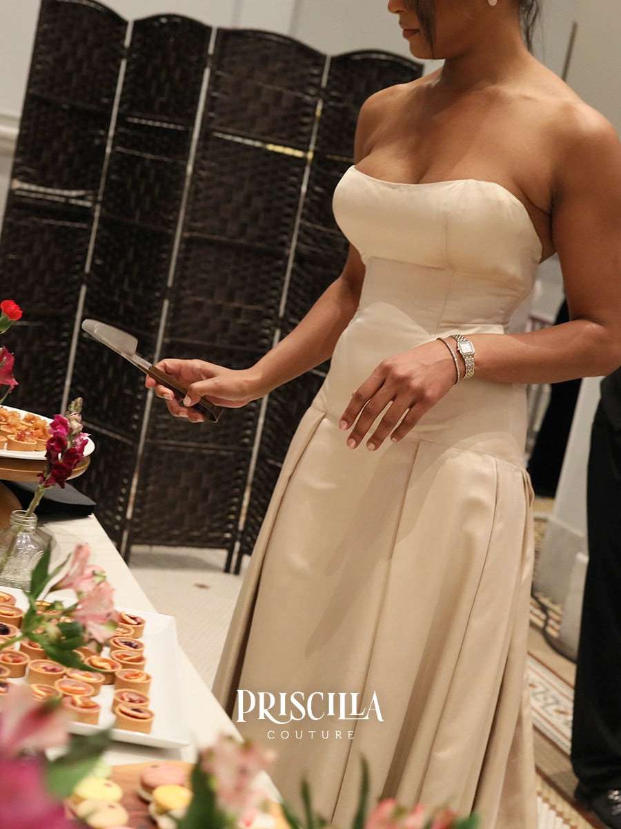 A woman in a strapless cream satin gown stands at a buffet table, holding a knife. The background features a dark wicker folding screen, and the brand name 'PRISCILLA COUTURE' is watermarked at the bottom.