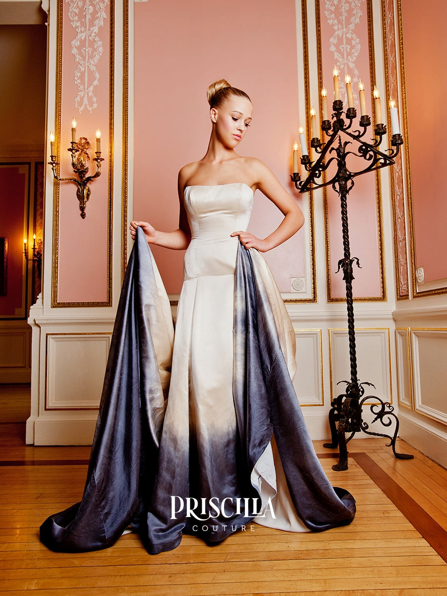 A woman models a strapless gown in an elegant room with pink walls, gold trim, and a large black candelabra. The couture wedding dress has a cream bodice and skirt with a dramatic metal gun grey ombré satin train.