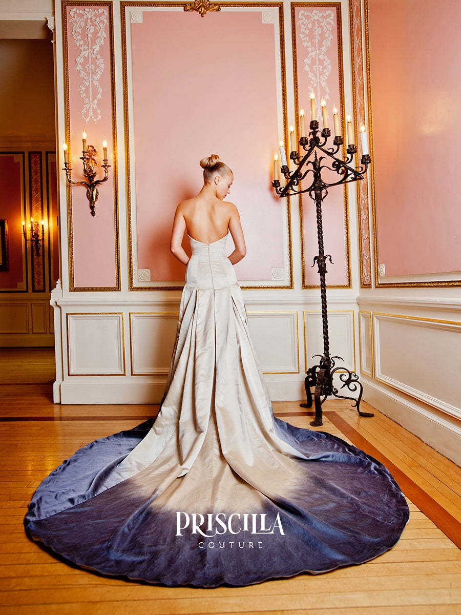A woman stands with her back to the camera wearing a Priscilla Couture bridal gown with a long dyed train. The dress has a cream upper bodice and skirt that fades into a deep metal gun grey at the hem, set in an elegant room with pink walls, gold trim, and a black candelabra.