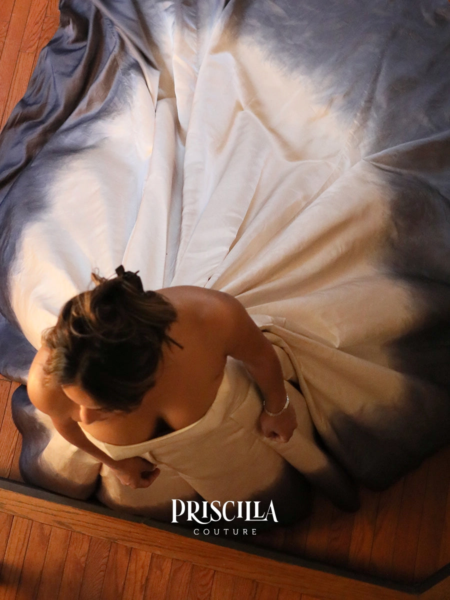 A woman from above wearing a voluminous champaign silk duchesse wedding gown with a dramatic gray-blue ombre effect near the edges, standing on a wooden floor. The brand name 'PRISCILLA COUTURE' is printed on the image.