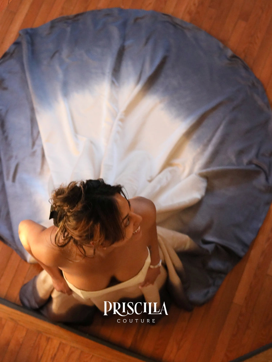 A woman stands on a polished wooden floor, wearing a champaign strapless couture wedding dress while looking down at a large circular blue and white tie-dye fabric spread out behind her. Overlay of the brand name 'PRISCILLA COUTURE'.