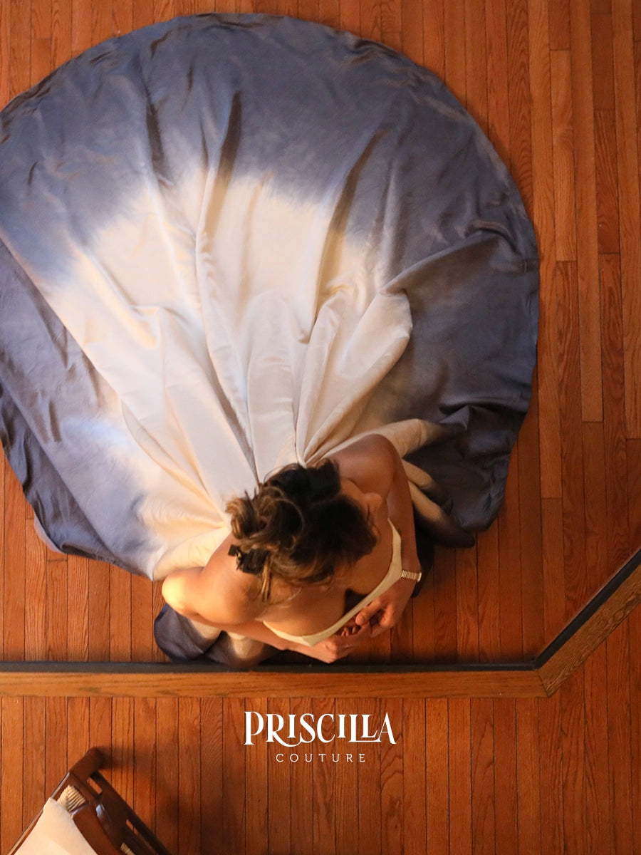 Overhead view of a woman wearing a voluminous metal gun grey and champaign dyed gown, standing on a wooden floor. The brand name 'PRISCILLA COUTURE' overlay up on pic.