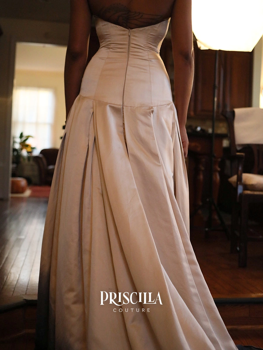 A woman wearing a Priscilla Couture champagne-colored silk duchesse gown, shown from the back in a dimly lit indoor setting with wooden floors and furniture.