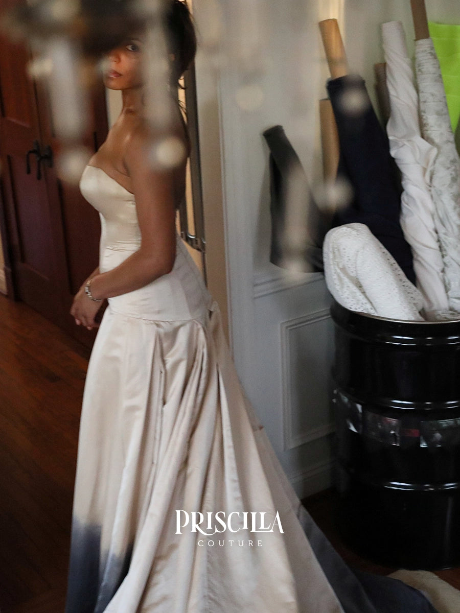 A woman wearing a strapless ivory silk duchesse wedding gown with a gradient fade to metal gun grey at the hem, standing in a room with a mirror and fabric rolls in the background. The brand name 'PRISCILLA COUTURE' is watermarked at the bottom.
