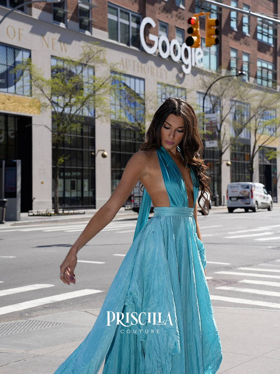 A woman wearing a flowing Priscilla Couture pool blue halter-neck maxi dress walks across a city street crosswalk. The dress has a deep V-neckline and a sheer, layered skirt.