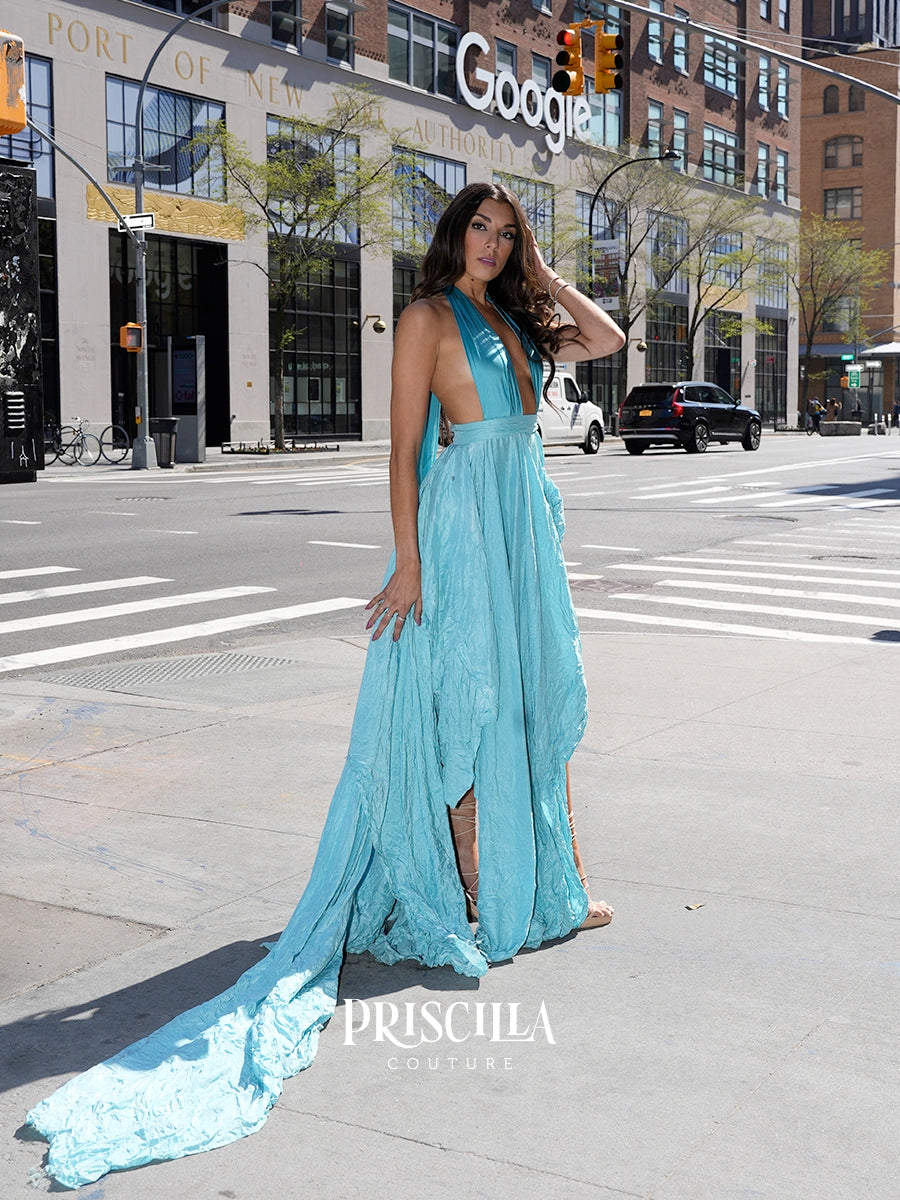 A woman models a light blue halter-neck maxi dress with a dramatic train on a city street. The dress features a deep V-neckline, side slit, and textured fabric.