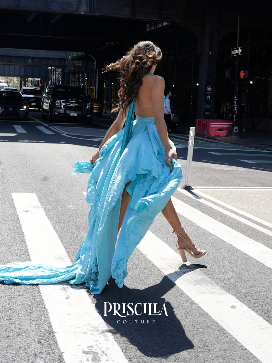 A woman walks across a New York City crosswalk wearing a light blue backless gown with a long flowing train and high slit. The brand name PRISCILLA COUTURE is displayed at the bottom of the image.