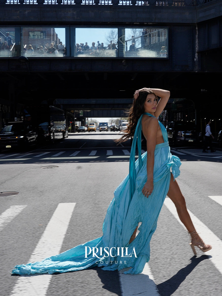 A model wearing a light blue halter gown with a long train walks across a city crosswalk under an elevated train track. The brand name 'PRISCILLA COUTURE' is displayed at the bottom.