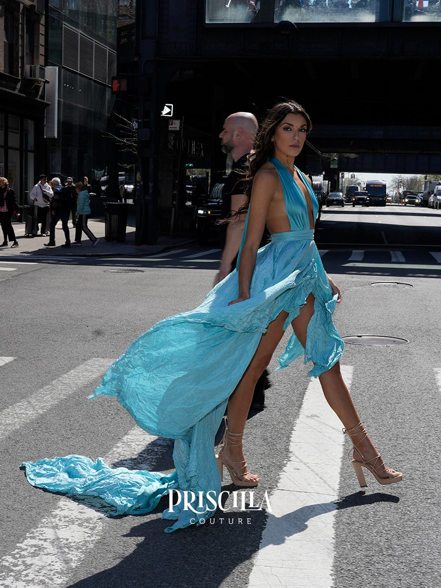 A woman walks across a New York City street wearing a flowing Priscilla Couture light blue halter gown with a long train and nude high-heeled sandals. A man is partially visible behind her.