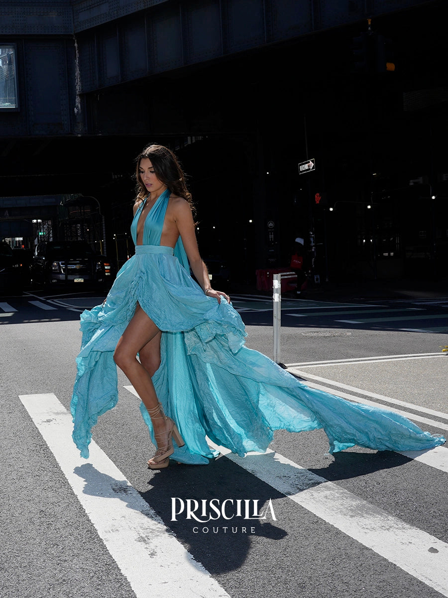 A woman models a flowing light blue halter-neck gown with a high leg slit and long draped train on a NYC crosswalk. The Priscilla Couture logo is visible at the bottom.