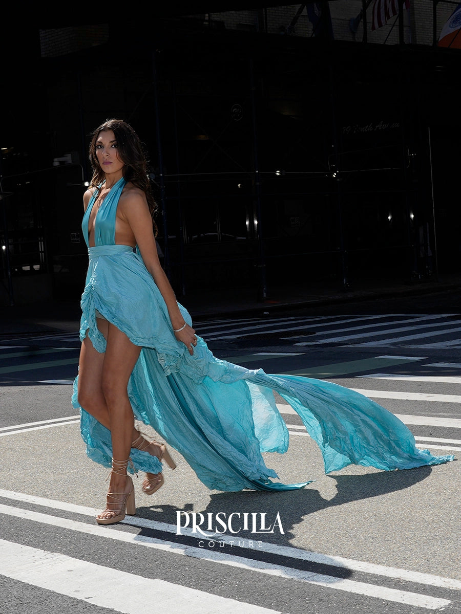 A woman walking across a NYC crosswalk wearing a flowing light blue halter dress with a high slit and long trailing skirt. The Priscilla Couture logo is visible at the bottom.