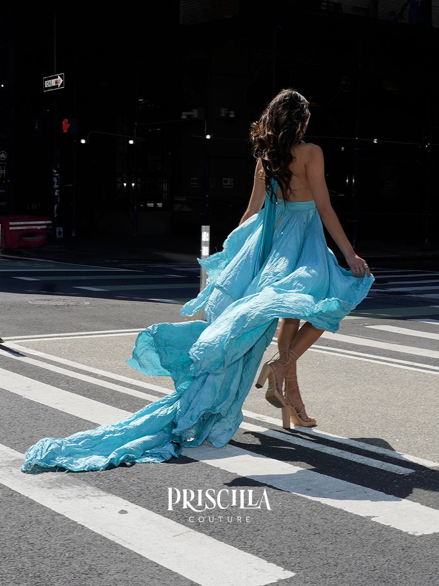 A woman wearing a flowing Priscilla Couture strapless light blue dress walks across a city crosswalk, photographed from behind. The dress has a long train that drapes across the street.