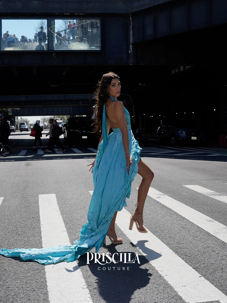 A woman models a light blue draped gown with a long train on a city crosswalk under an overpass. The brand name 'PRISCILLA COUTURE' is displayed at the bottom.