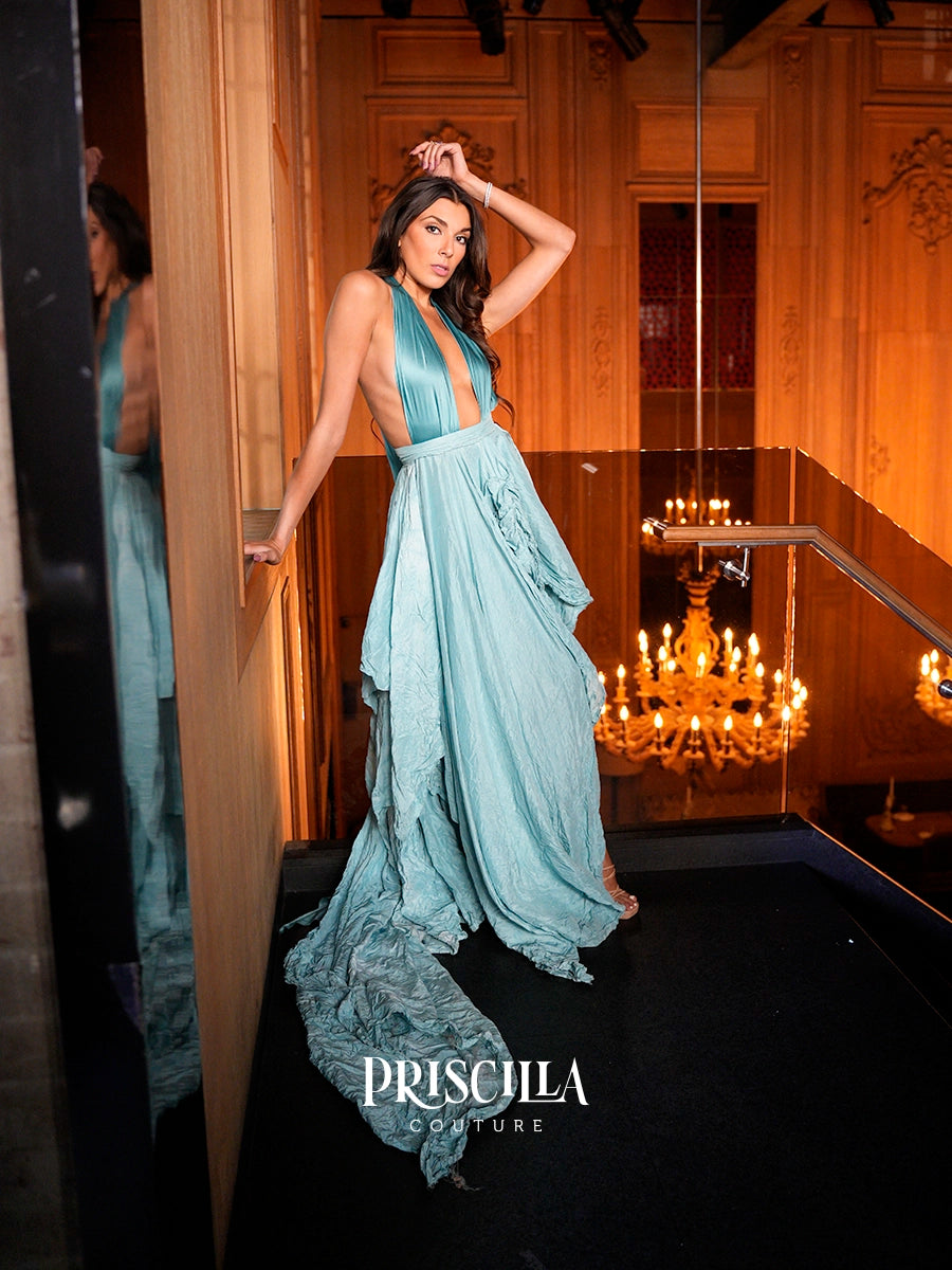 A woman models a light blue halter-neck evening gown with a deep V-neckline and a flowing, tiered skirt in an ornate, wood-paneled room with warm lighting and a chandelier. The brand name 'PRISCILLA COUTURE' is displayed at the bottom.