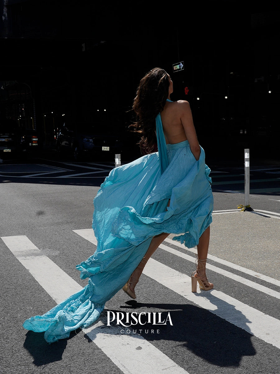 A woman wearing a flowing light blue one-shoulder dress walks across a city crosswalk at sunset, with the dress billowing in the wind. The brand name 'PRISCILLA COUTURE' is displayed at the bottom.