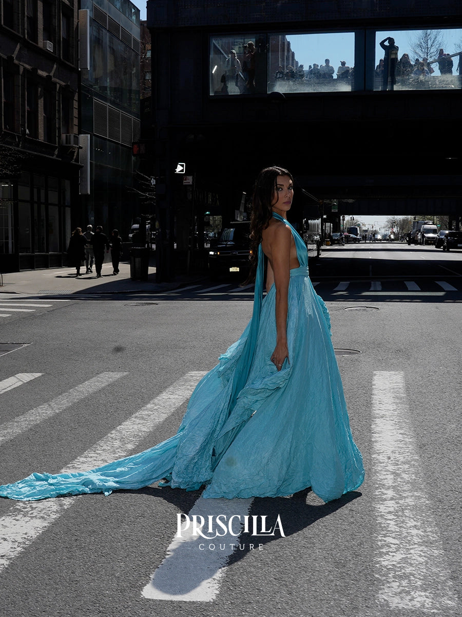 A woman stands on a city crosswalk wearing a flowing light blue evening gown with a long train. The photo is taken outdoors in an urban street setting under a bridge, with the brand name PRISCILLA COUTURE visible at the bottom.
