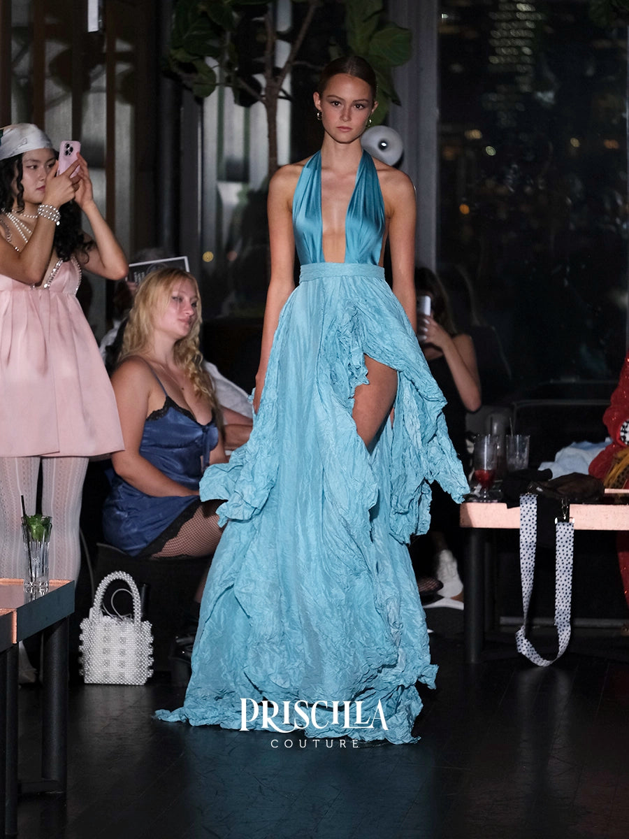 A model walks a fashion runway wearing a light blue halter-neck gown with a deep plunging neckline, a high-low ruffled hem, and a thigh-high slit. The PRISCILLA COUTURE logo is visible at the bottom of the image.