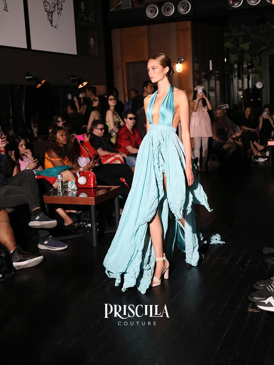 A model walks the runway wearing a light blue halter-neck gown with a high-low skirt and thigh-high slit. The Priscilla Couture logo is visible at the bottom of the runway.